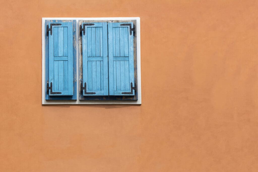 Blue wooden window shutters on the orange facade of an old house to illustrate Historic Metal Roofing Built to Last for Many Lifetimes